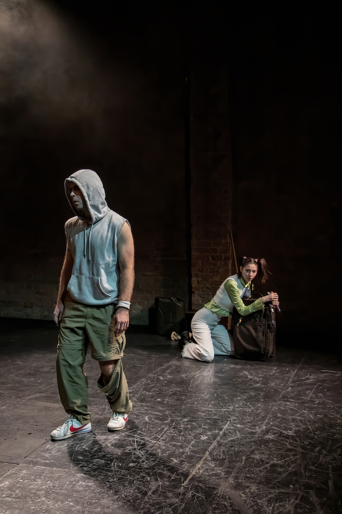 Actor (left) stands on stage wearing a gray hoodie with the sleeves cut off as a second actor crouches on her knees hugging a rolling suitcase behind him. 